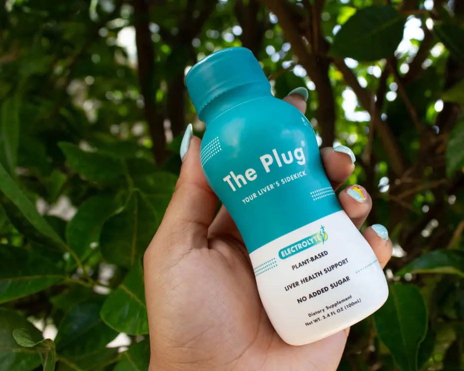A hand holds a bottle of The Plug Drink up against a backdrop of green leaves in the sun.