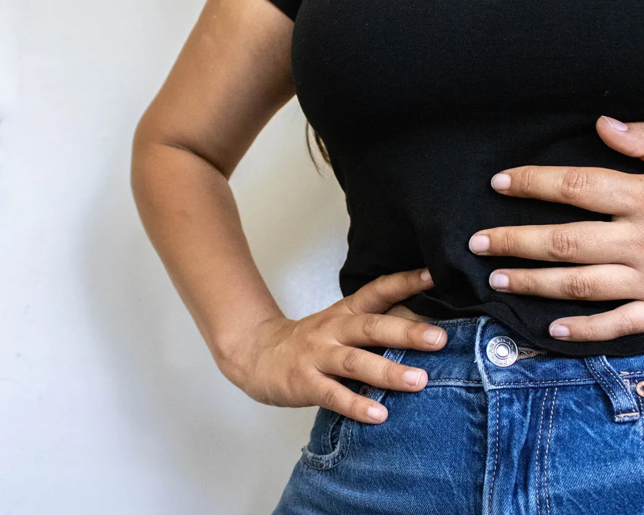 A woman in a black T-shirt and high-waisted jeans rests one hand on her stomach and the other on her hip.