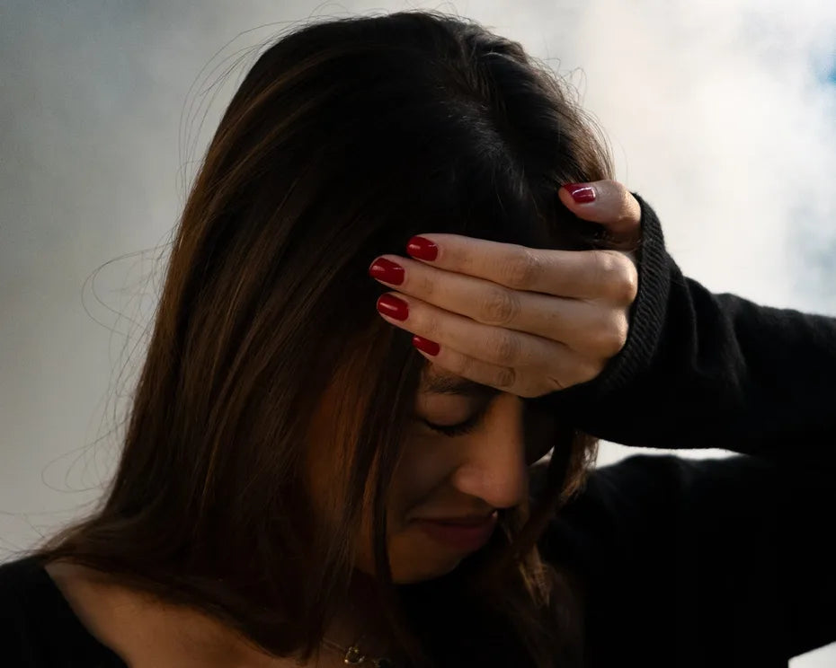 A woman with long dark hair presses her hand to her forehead and closes her eyes against a cloudy sky.
