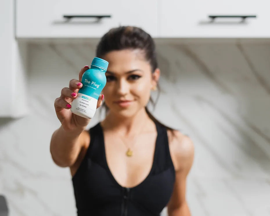 A smiling woman in a black sports top holds a bottle of The Plug Drink toward the camera in a bright white kitchen.