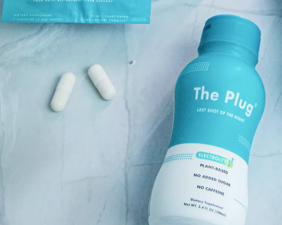 A bottle of The Plug Drink lies on a pale blue marble surface beside two white capsules and a packet of The Plug Pills.