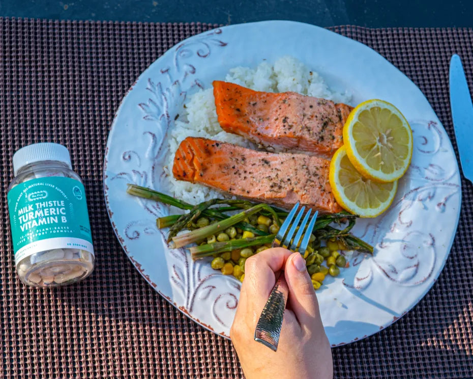 A hand holds a fork over a plate of salmon, rice, asparagus and lemon slices, with a bottle of The Plug Pills beside the plate.