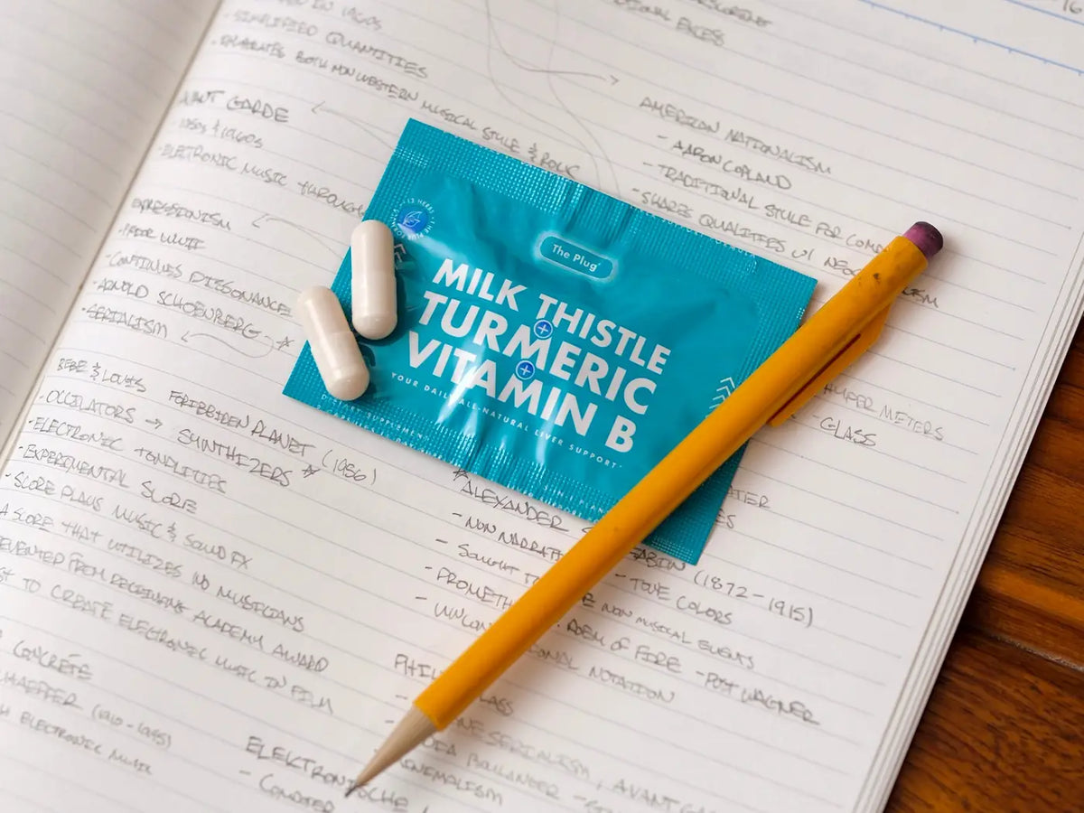 A packet of The Plug Pills and two white capsules rest on an open handwritten notebook beside a yellow pencil.