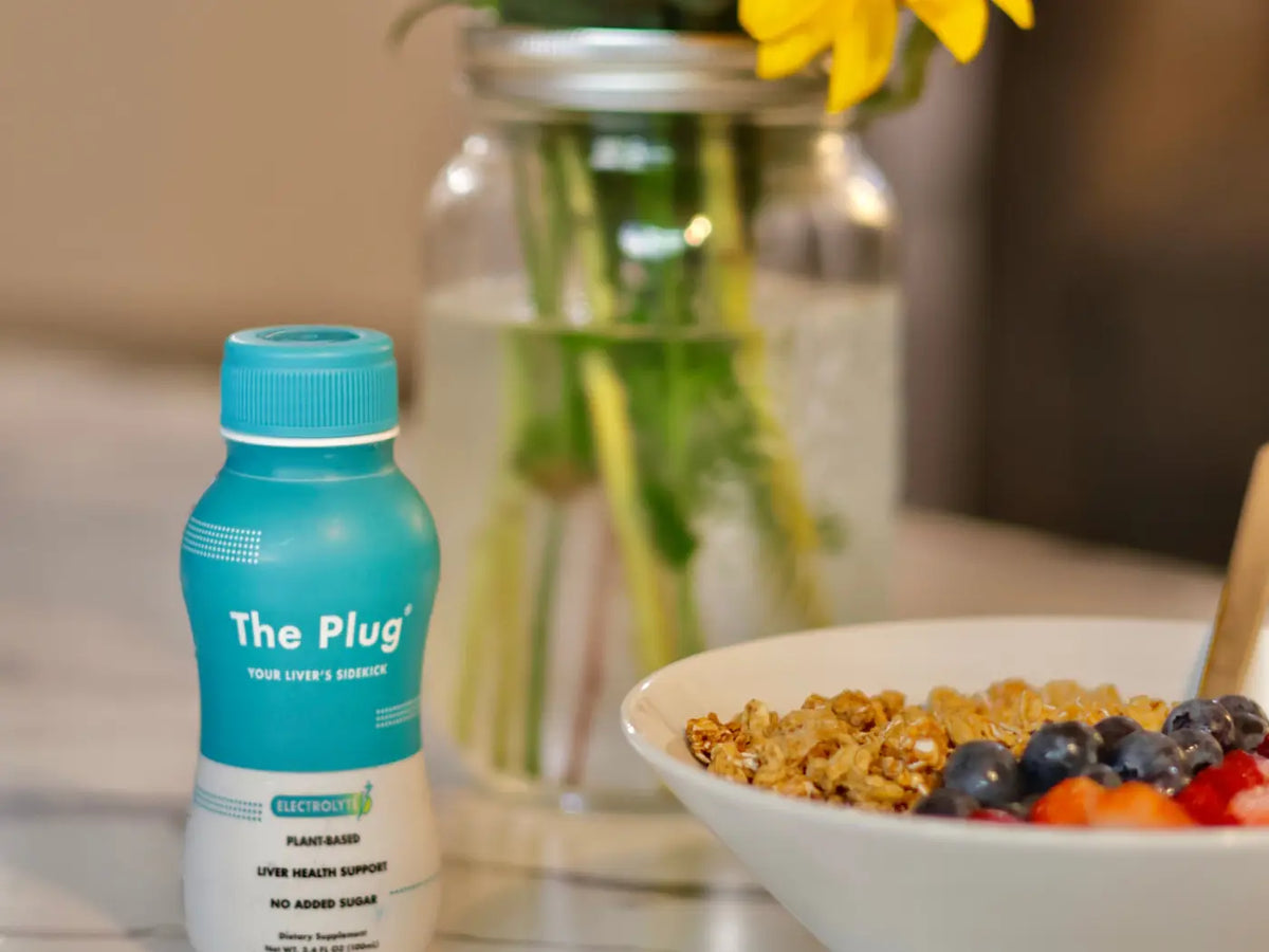 A bottle of The Plug Drink sits on a kitchen counter next to a bowl of granola with berries and a vase of sunflowers.
