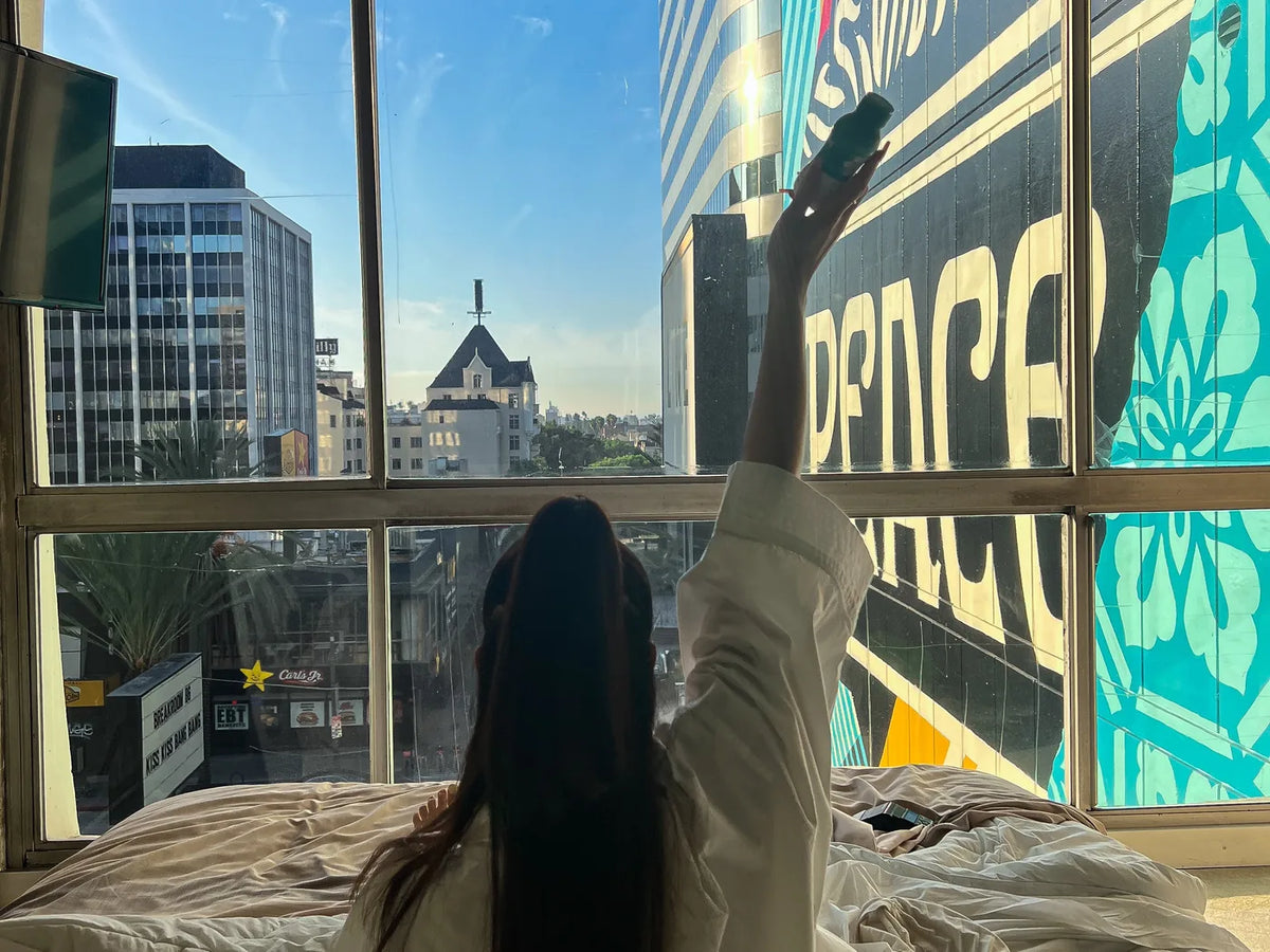 A woman sitting up in bed raises a bottle of The Plug Drink toward a sunny window overlooking city buildings.