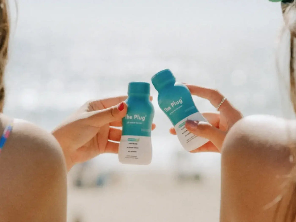 Two friends at the beach clink small bottles of The Plug Drink together in the sunshine.