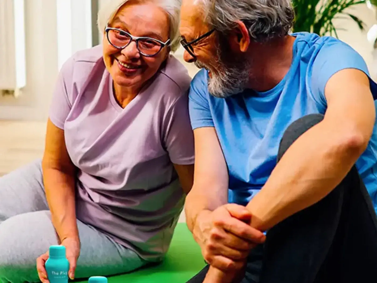 Two smiling older adults sit together on a green exercise mat at home with two bottles of The Plug Drink beside them.