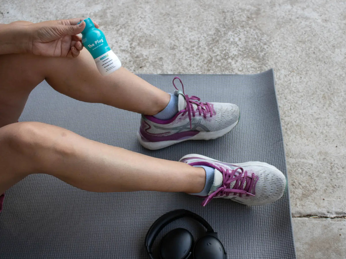A person in sneakers sits on a gray yoga mat holding a bottle of The Plug Drink, with headphones beside them.