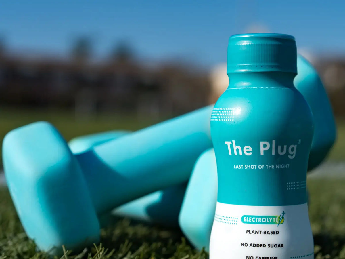 A bottle of The Plug Drink stands on sunlit grass in front of a pair of teal dumbbells.