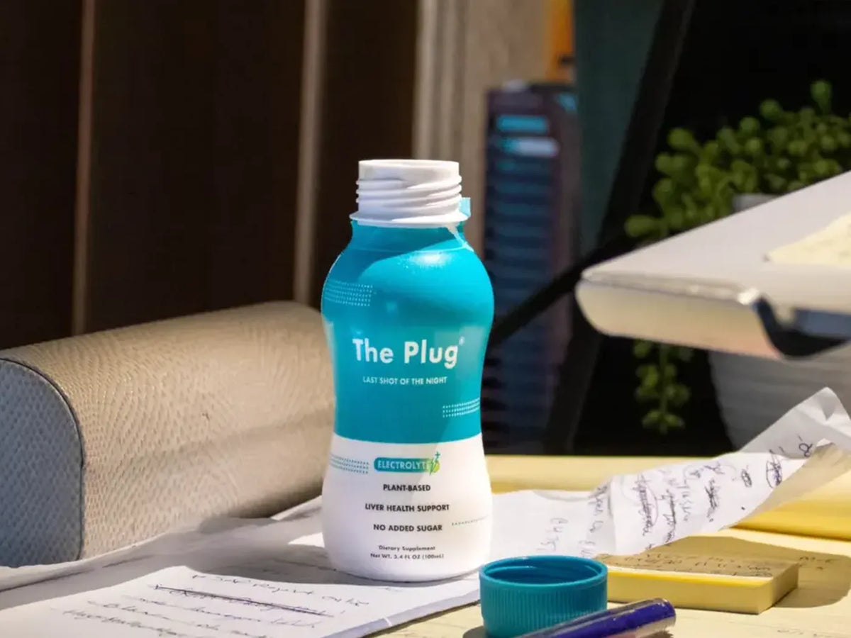 An open bottle of The Plug Drink stands on a sunny desk beside handwritten notes, a pen and a teal bottle cap.