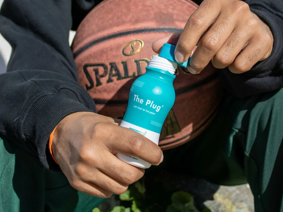 Two hands twist the cap off a bottle of The Plug Drink in front of a basketball.