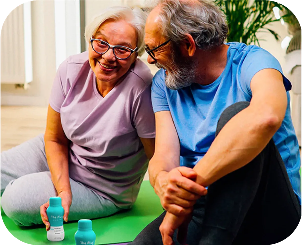 Older couple stretching on a yoga mat with two bottles of The Plug liver support drink