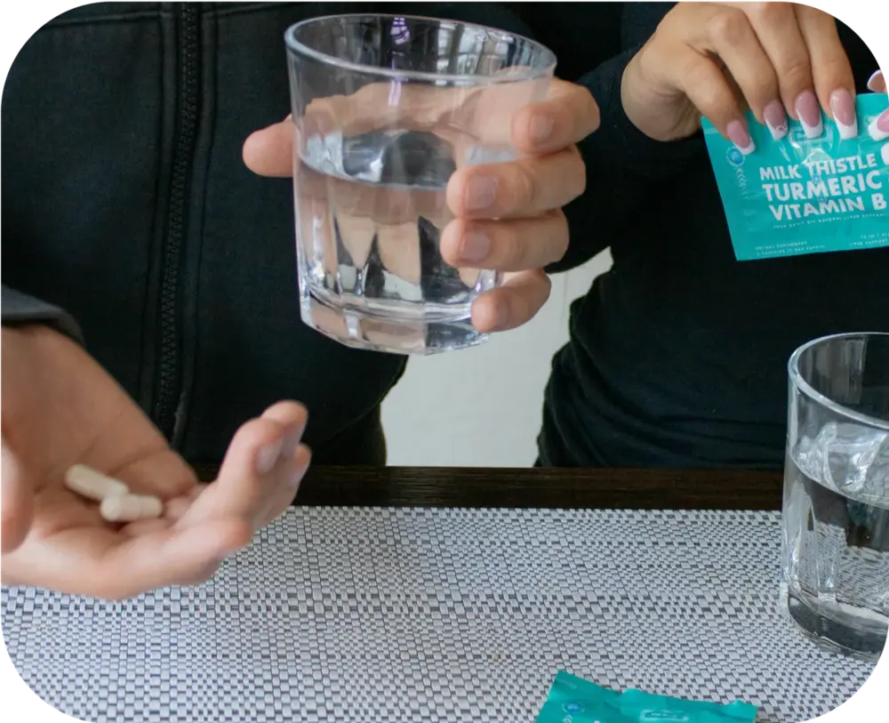 Person holding Milk Thistle Turmeric Vitamin B liver support capsules alongside a glass of water