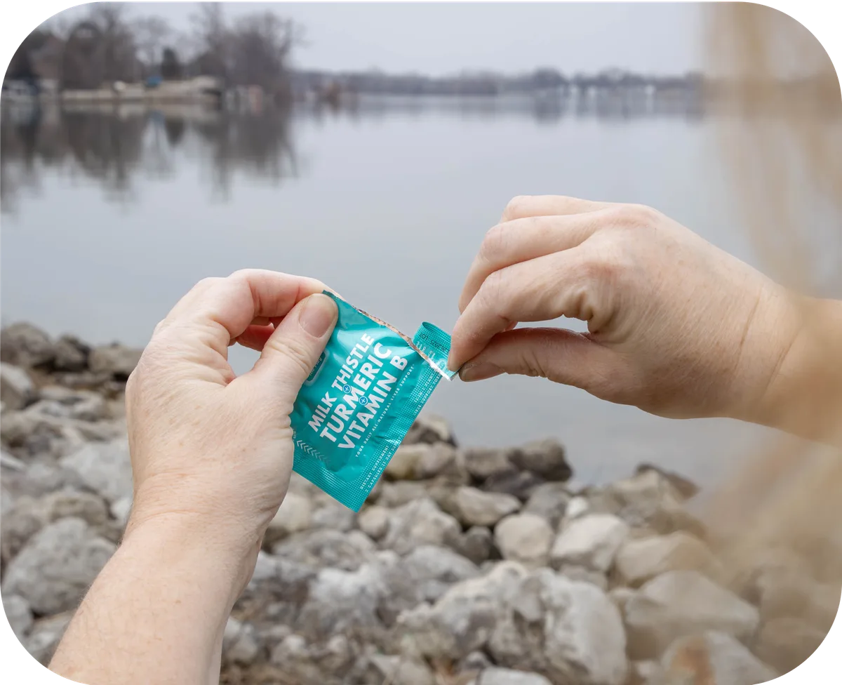 Person opening a Milk Thistle Turmeric Vitamin B liver support packet outdoors by a lake