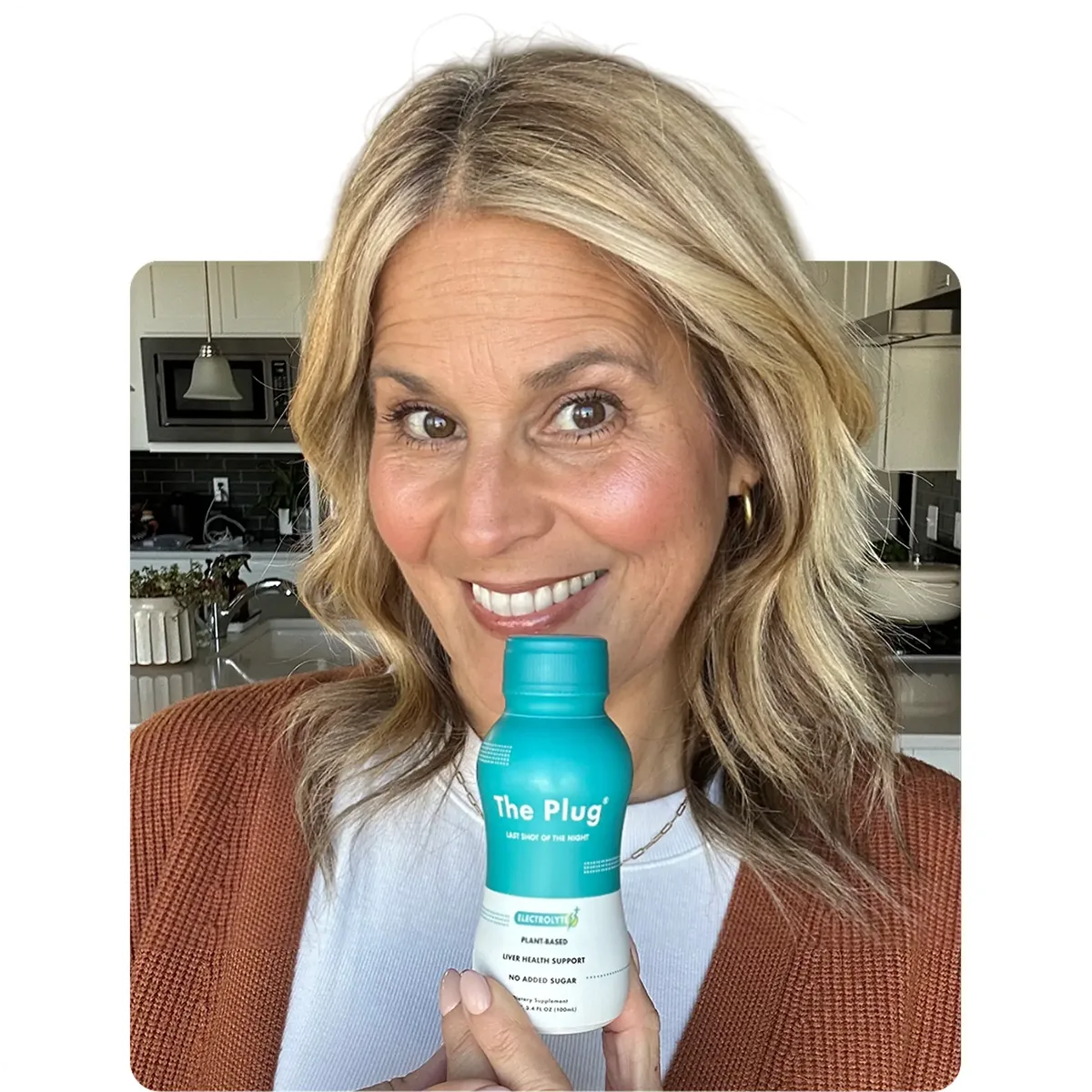 Smiling woman holding The Plug liver support drink bottle in her kitchen