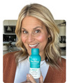Smiling woman holding The Plug liver support drink bottle in her kitchen
