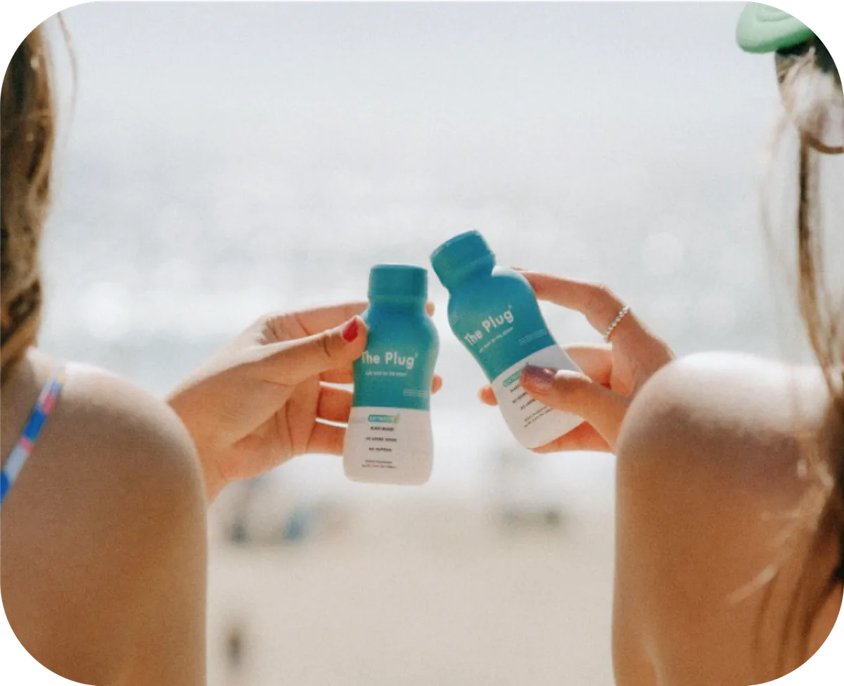 Two hands clinking bottles of The Plug liver support drink on the beach
