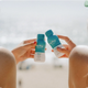 Two hands clinking bottles of The Plug liver support drink on the beach