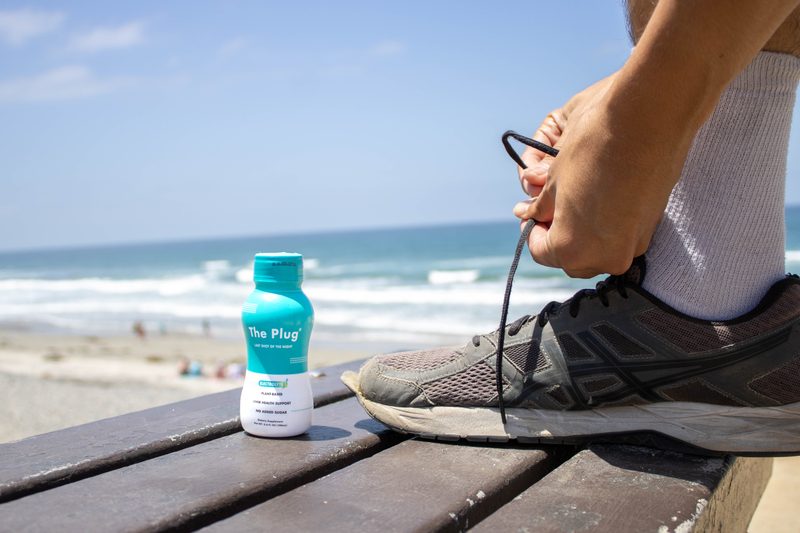 A bottle of The Plug Drink on a beach bench while a runner ties a shoe